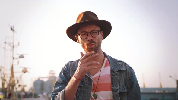 Portrait of Stylish Hipster Bearded Man in Hat and Glasses Smoking Cigarette Outdoors alt