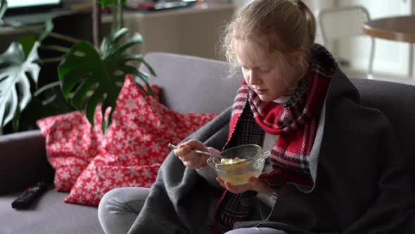 Portrait of a Sick Child in a Scarf and Plaid, Girl Eating Broth on the Sofa in the Apartment