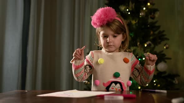 New Year. Little Pretty Girl Sits At The Table And Draws On Piece Of Paper Drawing. alt