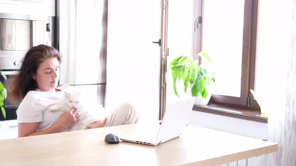 A Female Freelancer is Sitting in an Armchair at a Table with Plants Remotely Working on a Laptop in