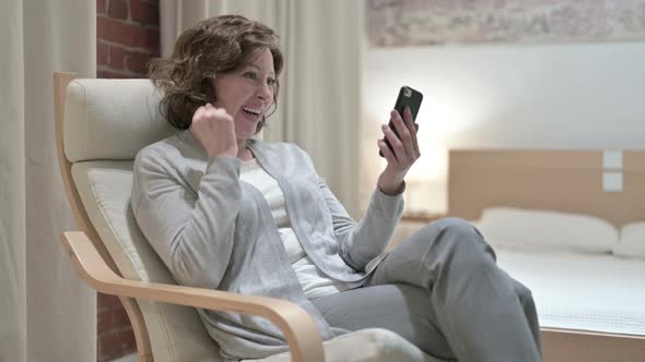 Old Woman Celebrating Success on Smartphone on Sofa  alt