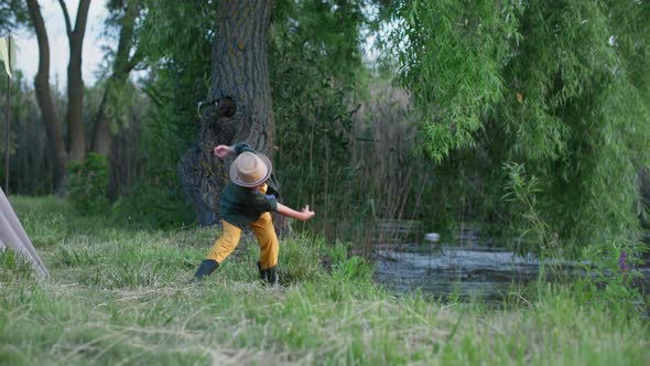 Joyful Childhood Happy Boy in Rubber Boots and Hat Enjoys Relaxing in Nature and Throws Stones Into alt