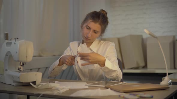 Serious Brunette Tailor Cutting Covid-19 Face Mask with Scissors, Sighing, and Holding Head in Hands alt