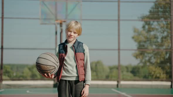 Boy Stands on a Court Holding Basketball Ball and Looking at the Camera alt