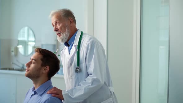 Mature Male Doctor Chiropractor Osteopath Talking with Young Man Patient Sitting in Chair with alt