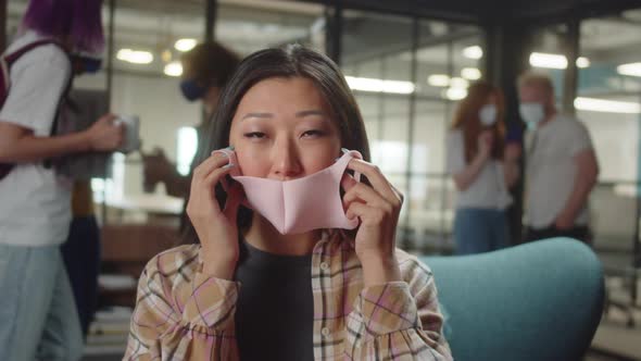 Sitting in the Entrance Hall in the University a Young Asian Lady is Putting a Protective Mask on alt