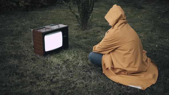 Man in a Yellow Raincoat is Sitting on Grass and Looking at Old Retro TV