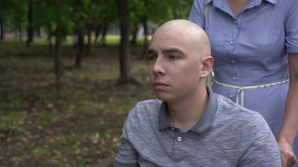 A Woman Is Driving a Young Man with Oncology in a Wheelchair Through the Park Closeup. The Man Is alt