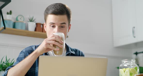 Young Man Works or Studies Online on Laptop in Kitchen, Drinks Coffee in Morning