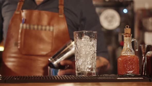 Bar Rack with Tall Glass with Ice Cubes Inside Barman Is Moving in Background alt