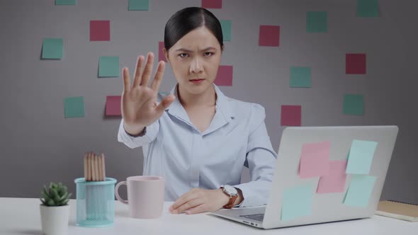 Asian woman working on a laptop looking at camera showing hand making stop sign