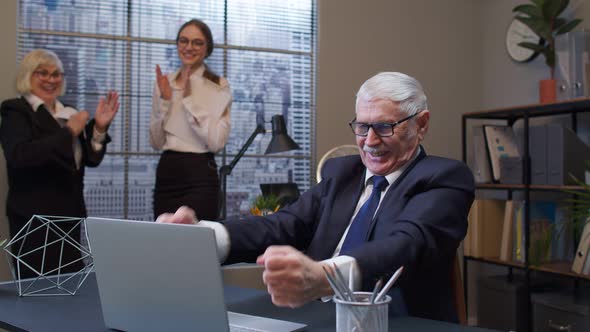 Cheerful Mature Old Businessman Raising Hands Celebrating Sudden Victory with Colleagues in Office alt