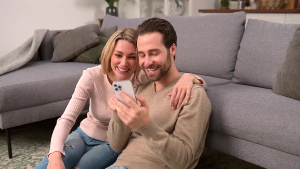 Couple in Love are Sitting on the Floor at Home and Using Smartphone alt