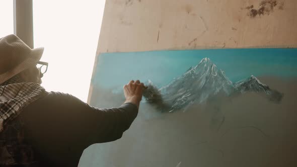 Old Artist Is Drawing By White Pastel Mountains on a Canvas alt