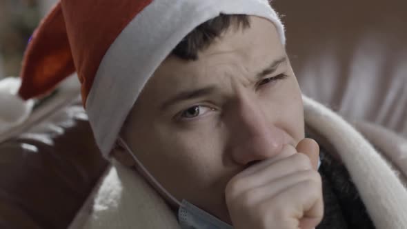 Closeup Headshot of Young Ill Man in Christmas Hat Coughing and Looking at Camera alt