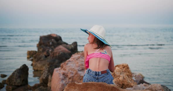 Teen Girl in Beach Clothes and Hat is Sitting on Stone on Seashore Rear View