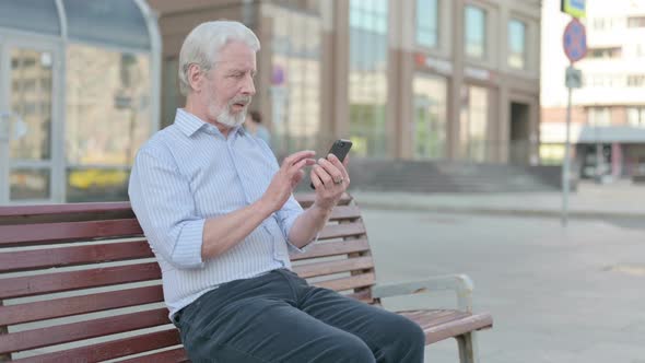 Old Man Celebrating Online Success on Smartphone While Sitting Outdoor on Bench alt