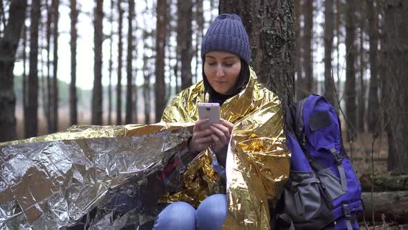Positive Woman Tourist Wrapped in a Golden Forest Survival Blanket Uses a Smartphone alt