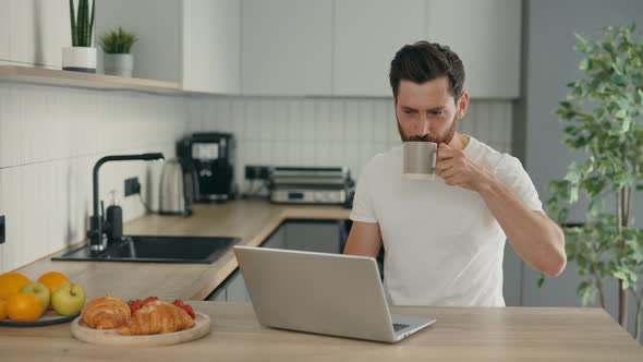 Handsome Man is in His New Home Sitting on the Chair in Kitchen Using Laptop for Distance Work and alt