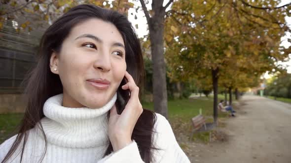 Asian Pretty Girl Wearing White Sweater Talking By Smartphone and Walking in Park.