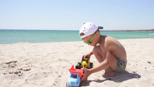 Boy Dressed in Sunglasses and a Baseball Cap Plays Cars on a Sandy Beach Against the Beautiful Sea alt