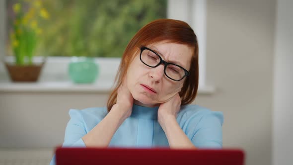 Front View Overworking Caucasian Mature Woman Stretching Neck Massaging Sitting at Table with Laptop alt