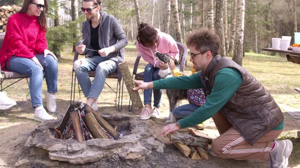 Happy Friends Having Barbecue Party in Nature, Young Man Adds Wood To Fire