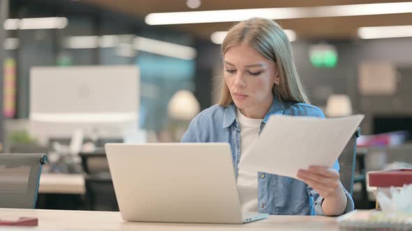 Successful Young Woman Reading Documents While Using Laptop alt