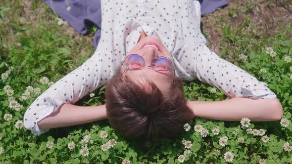 Top View on Woman with Colorful Sunglasses Lying on Lawn in Urban Park and Enjoys Flowers alt