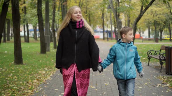 Portrait of Caucasian Woman and Boy Walking in Autumn Park Holding Hands Talking and Leaving alt