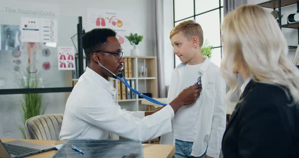 Male Doctor Checking Boy's Lungs During His Scheduled Visit to Clinic with Beautiful mother alt