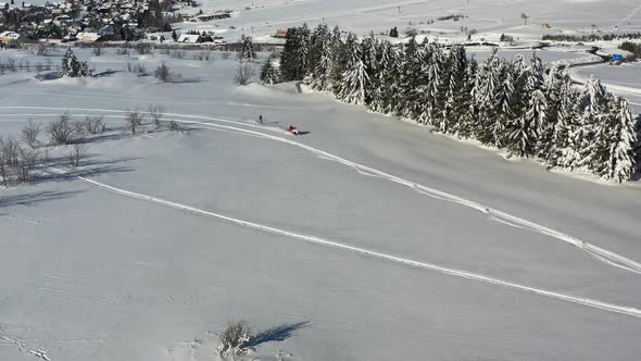 Drone aerial view of winter fun on snow, snowmobile towing skier on flat ground on sunny day alt