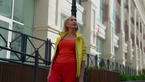a Blonde in Red Clothes and a Yellow Jacket Leaned Against a Pole on a City Street alt