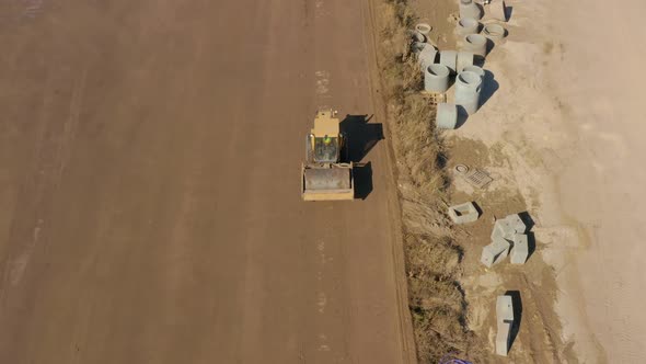 Aerial following shot of a Bulldozer roller compacting dirt in a field next to drain pipes alt