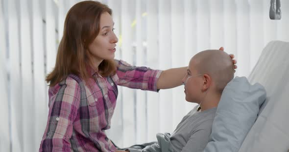 Side View of Woman Talking To Sick Son Resting in Bed After Chemotherapy in Hospital alt