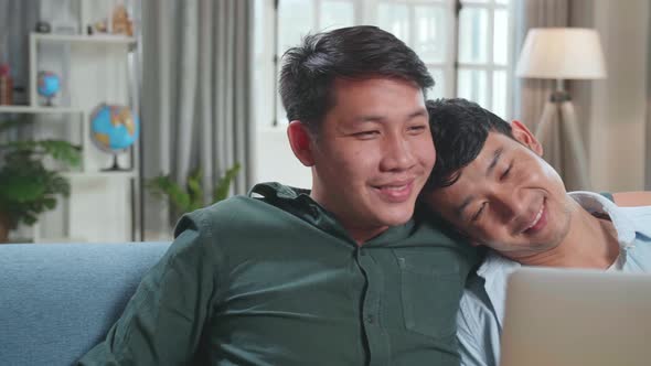 Asian Gentle Gay Couple Using Laptop Computer While Sitting On A Couch In Apartment alt