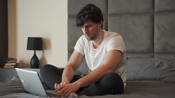Young Smiling Man in a White Tshirt is Sitting in Bed Working on a Laptop Freelancing at Home alt