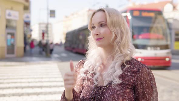 A Middleaged Caucasian Woman Shows a Thumb Up to the Camera and Nods with a Smile in an Urban Area alt