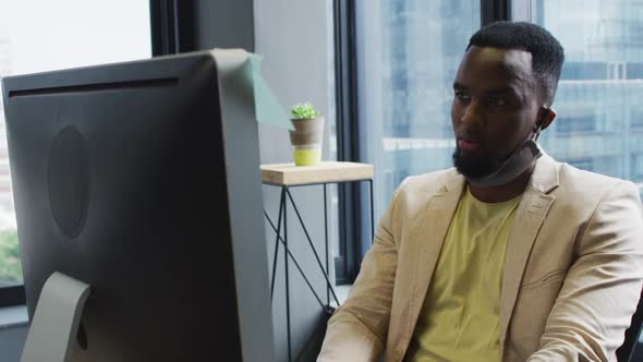 African american man with face mask around his neck using computer while sitting on his desk at mode alt