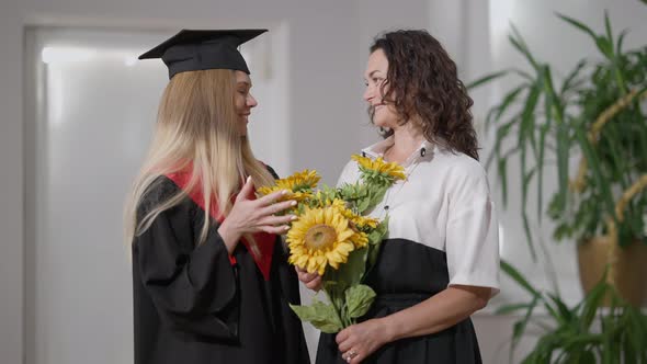 Happy Proud Mother and Daughter Graduate with Bouquet of Flowers Standing Indoors Smiling Looking at alt