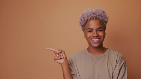 Dark Skinned Woman with Appealing Smile Pointing on Left Side Over Brown Wall alt