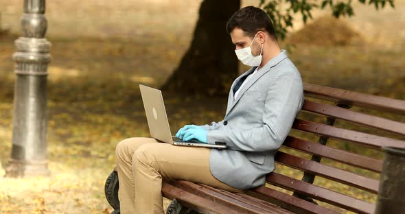 Young businessman with face mask working outdoors on bench in city, using laptop.