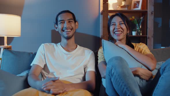asia couple man and woman smile and laugh lay down on sofa in living room at night watch comedy.