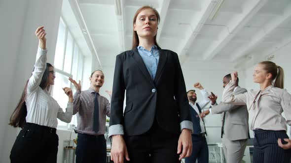Low Angle Shot of Ambitious Woman Standing in Office with Serious Face While Coworkers Dancing alt