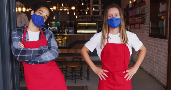 Two diverse male baristas wearing face masks and aprons standing in doorway of cafe alt