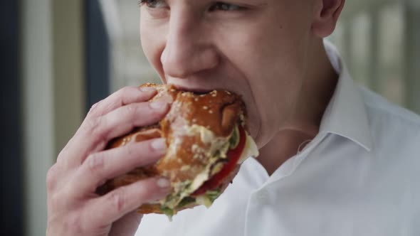 Close View of Handsome Man in Shirt Eating Tasty Hamburger Indoor at Window alt