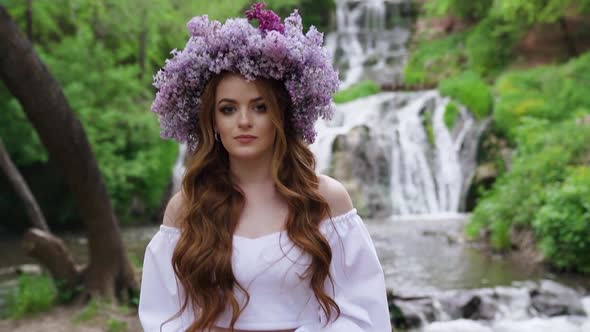 A young woman with lilac wreath walking down near waterfall in the background. alt