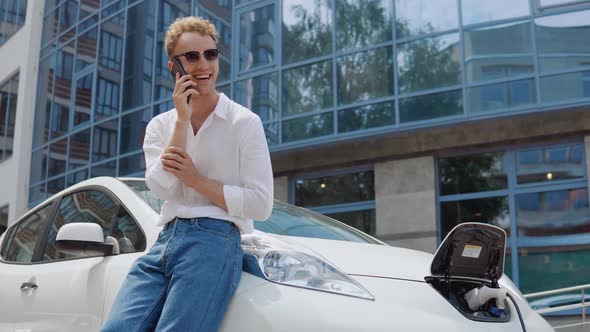 Stylish Modern Young Curly Man Charges His Electric Car in the Yard of a Residential Complex and alt