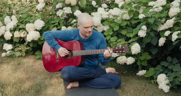 Caucasian Man is Sitting in a Park Among Flowers and Playing Red Acoustic Guitar