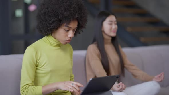 Concentrated Young Beautiful African American Woman Typing on Tablet Keyboard As Relaxed Asian Lady alt